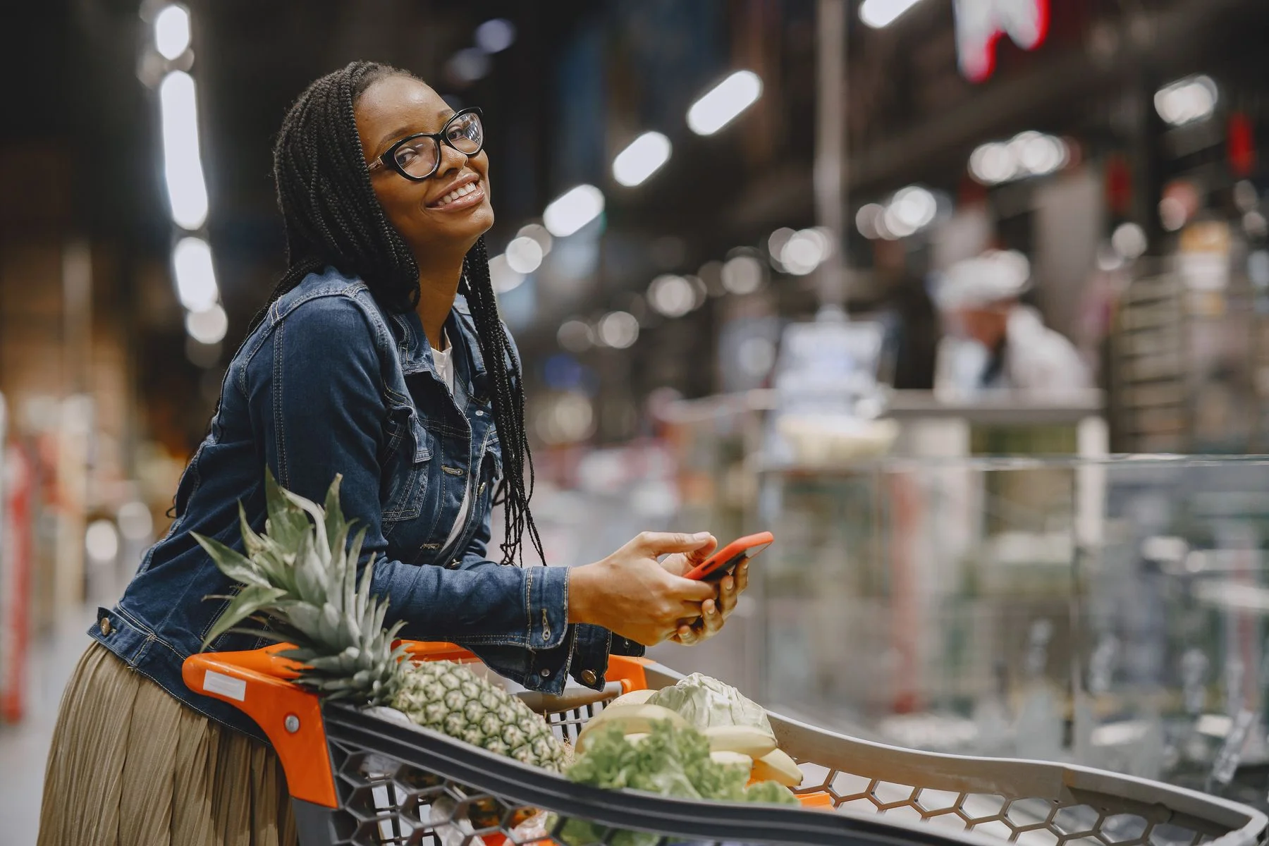 A woman smiling while holding her phone beside a shopping trolley